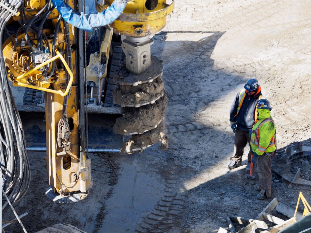 Two workers in protective gear at a construction site with heavy drilling machinery.